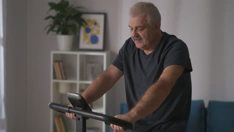 grey-haired-middle-aged-man-is-training-on-exercise-bike-at-home-caring-about-health-keeping-fit-in-old-age-sport-activity